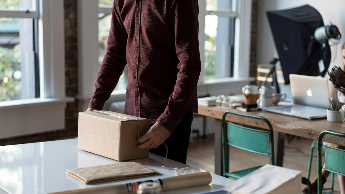 A person packing a cardboard parcel at a workspace beside a laptop