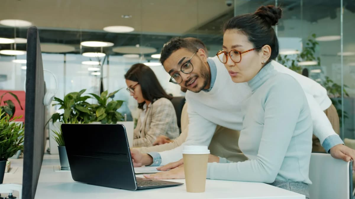 Two colleagues leaning over one laptop in a bright open-plan office