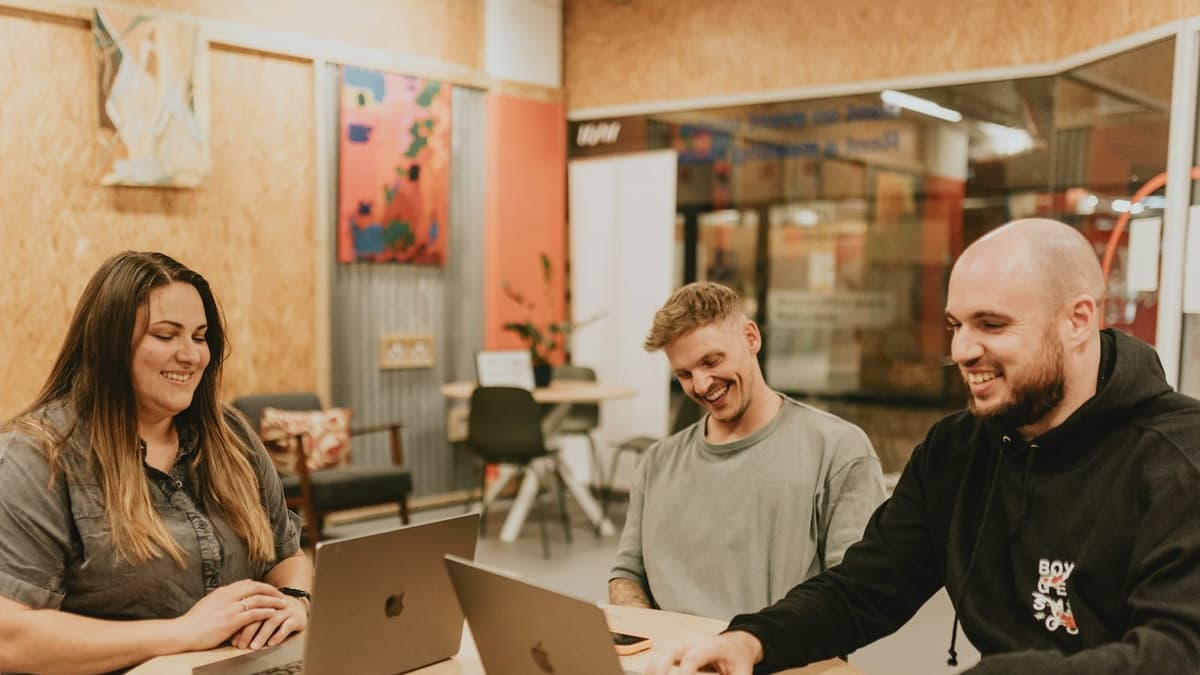 Three colleagues laughing while working together on laptops at a shared office table