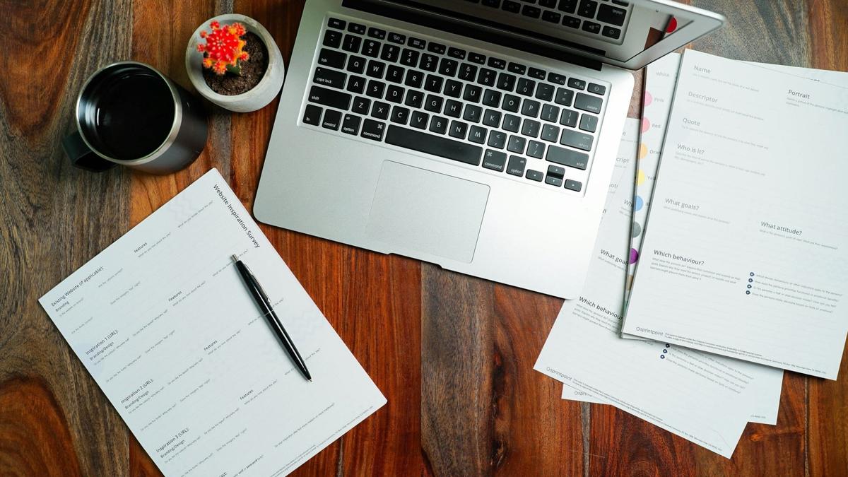 An overhead view of a laptop, business worksheets, pen and coffee on a wooden desk