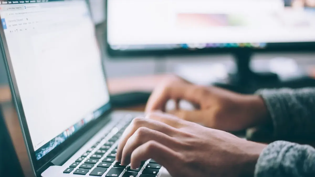 Close-up of hands typing on a laptop at a desk with a second monitor
