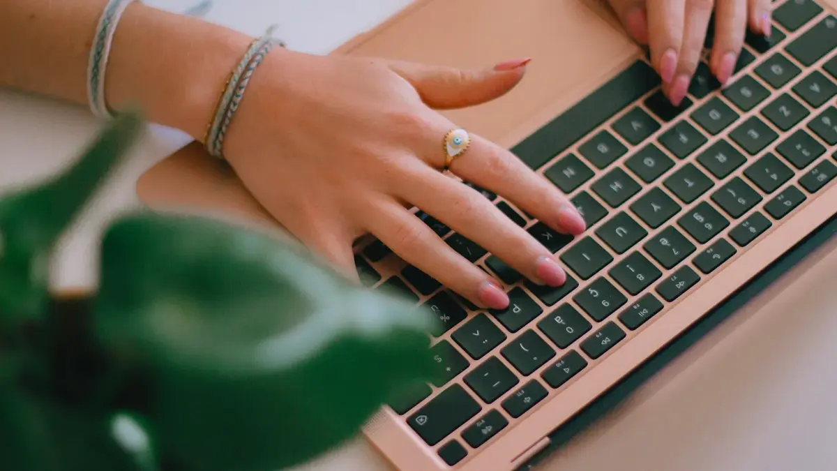 Close-up of a creator's hands typing on a rose-gold laptop keyboard beside a houseplant