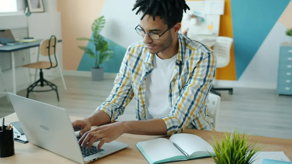 A writer in a checked shirt typing on a laptop next to an open notebook in a bright studio