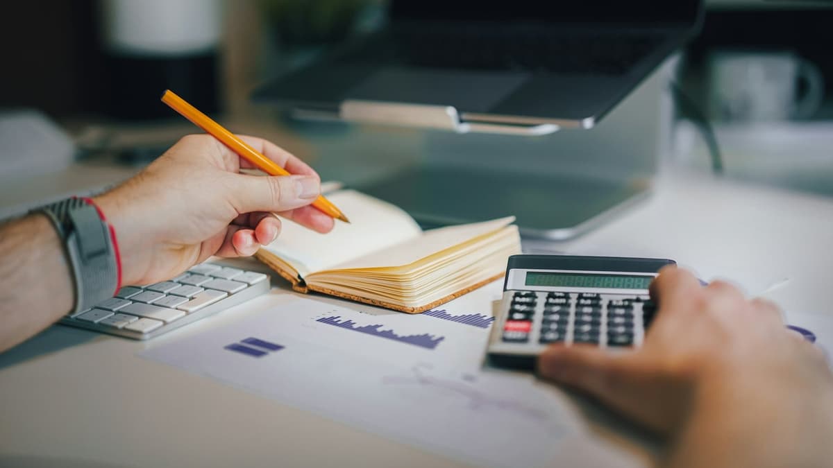Hands using a calculator and writing in a notebook beside printed charts, a keyboard and a laptop