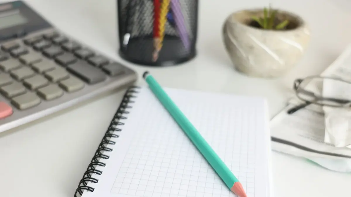 A green pencil across an open grid notebook beside a calculator, glasses and a small plant