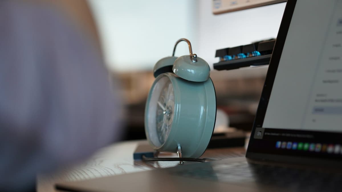 A pale blue twin-bell alarm clock on a wooden desk beside an open laptop and a keyboard