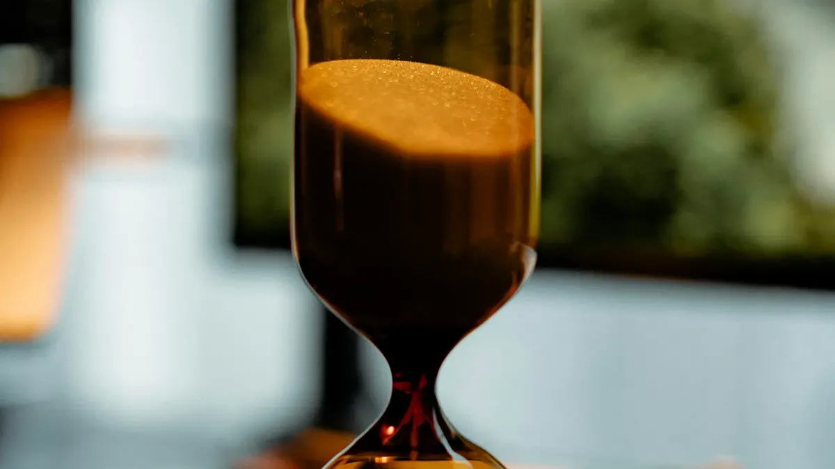An amber hourglass with sand running through it on a desk, with a blurred office window behind