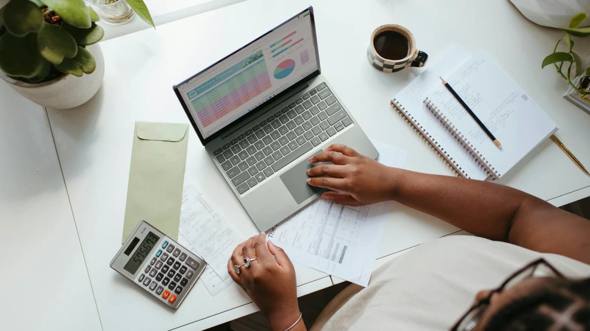 A person reviews papers beside a laptop, calculator and coffee on a desk
